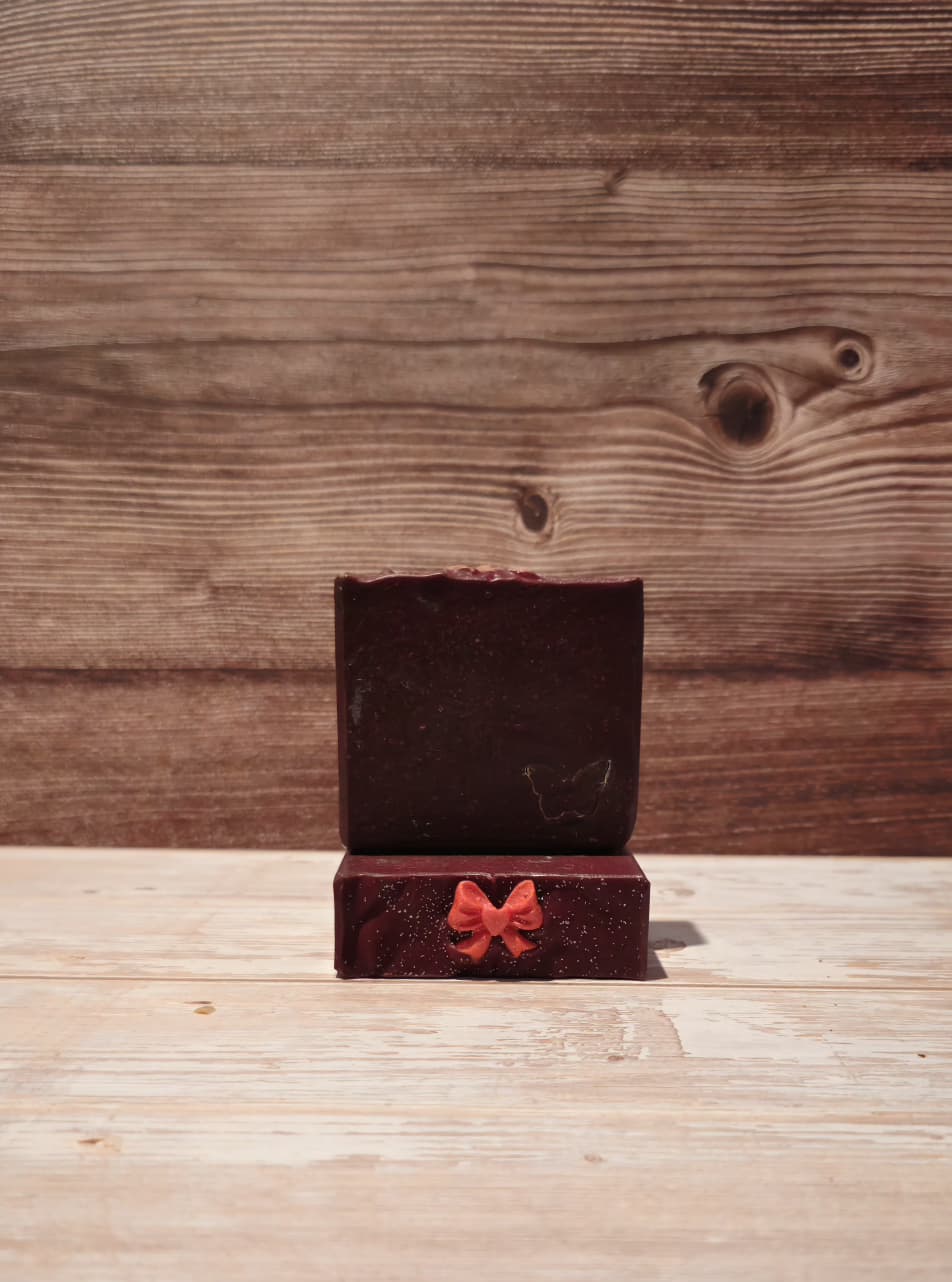 A dark red wax melt or soap block displayed in an open wooden gift box with a small red bow on the front, set against a rustic wooden plank background.