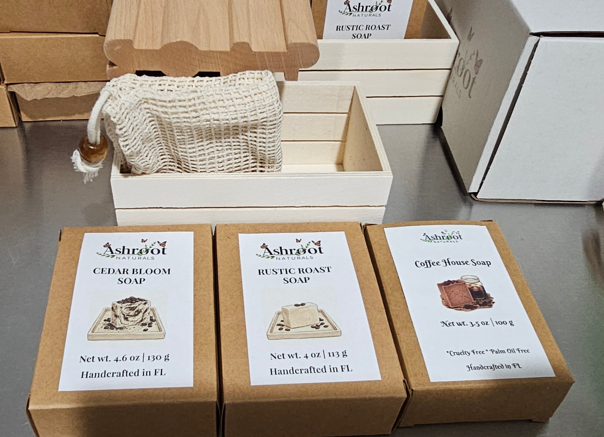 Three boxed handcrafted soaps are displayed on a stainless steel table, alongside a natural fiber soap bag and wooden boxes.