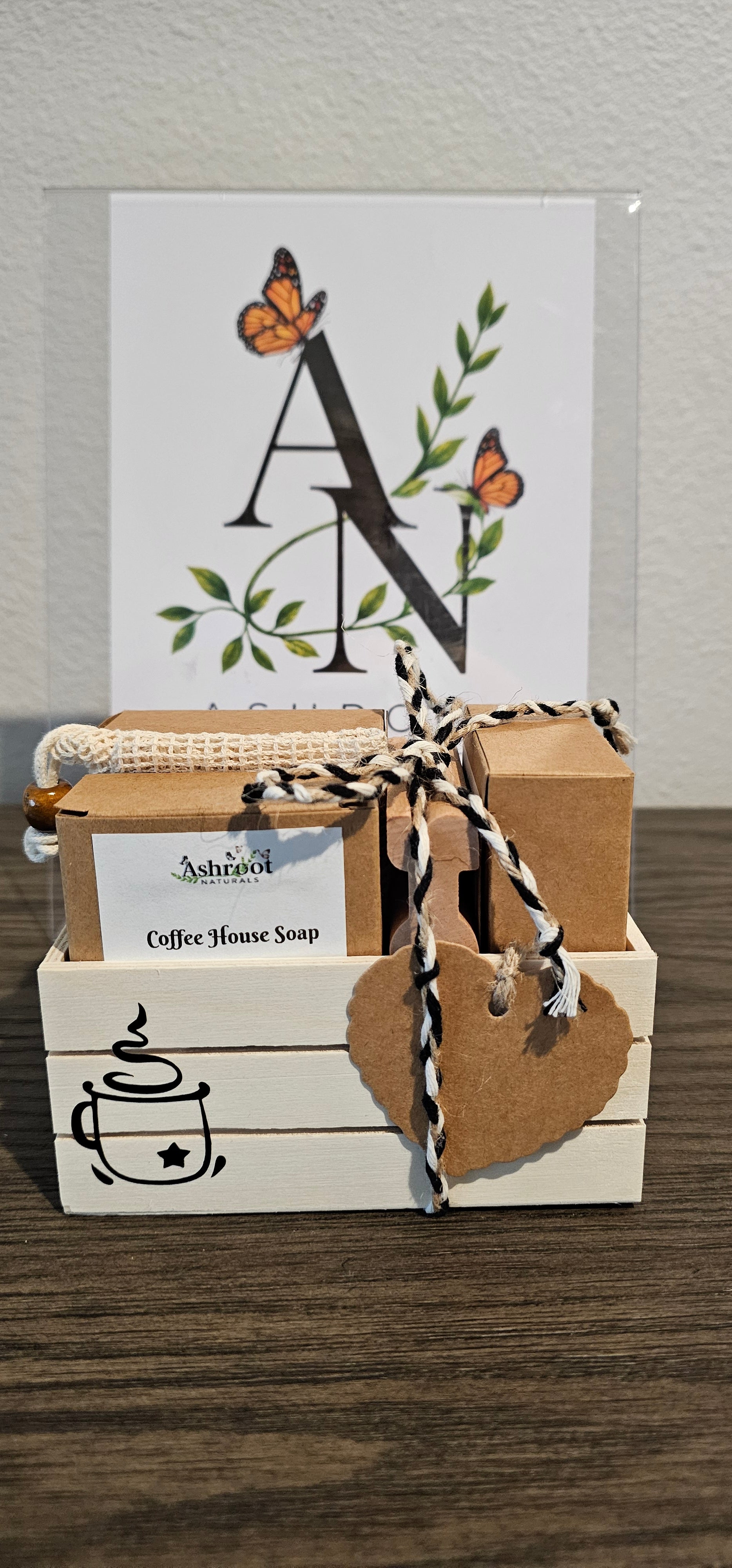 A small kraft paper gift box tied with black, kraft-and-white twine, placed next to a printed card featuring a large decorative letter A, butterflies, and green leaves logo image. The wood crate box has a heart kraft paper hanging tag.