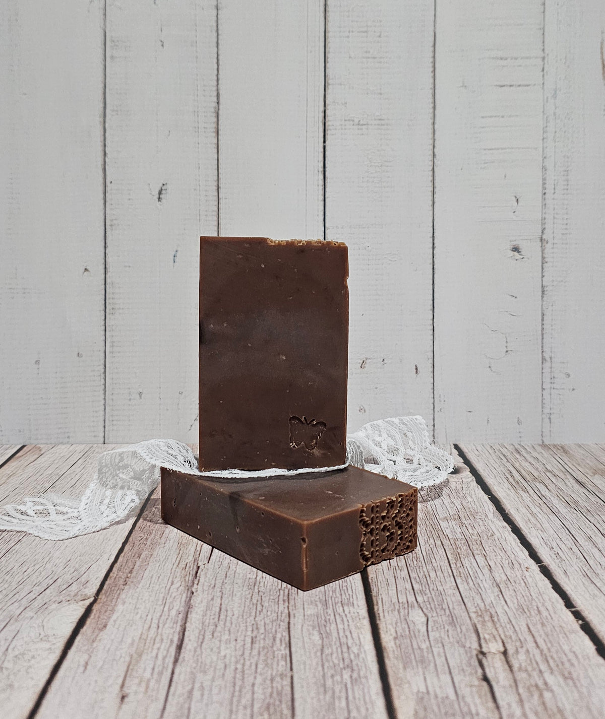 Two rectangular dark brown bars of soap standing upright on a rustic white wooden surface, with a small white lace ribbon tied around them.