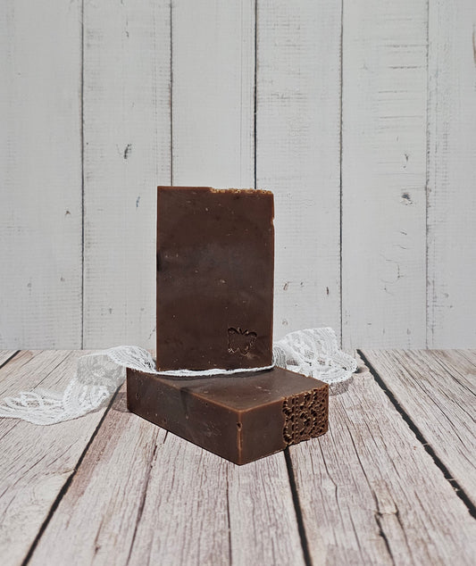 Two rectangular dark brown bars of soap standing upright on a rustic white wooden surface, with a small white lace ribbon tied around them.