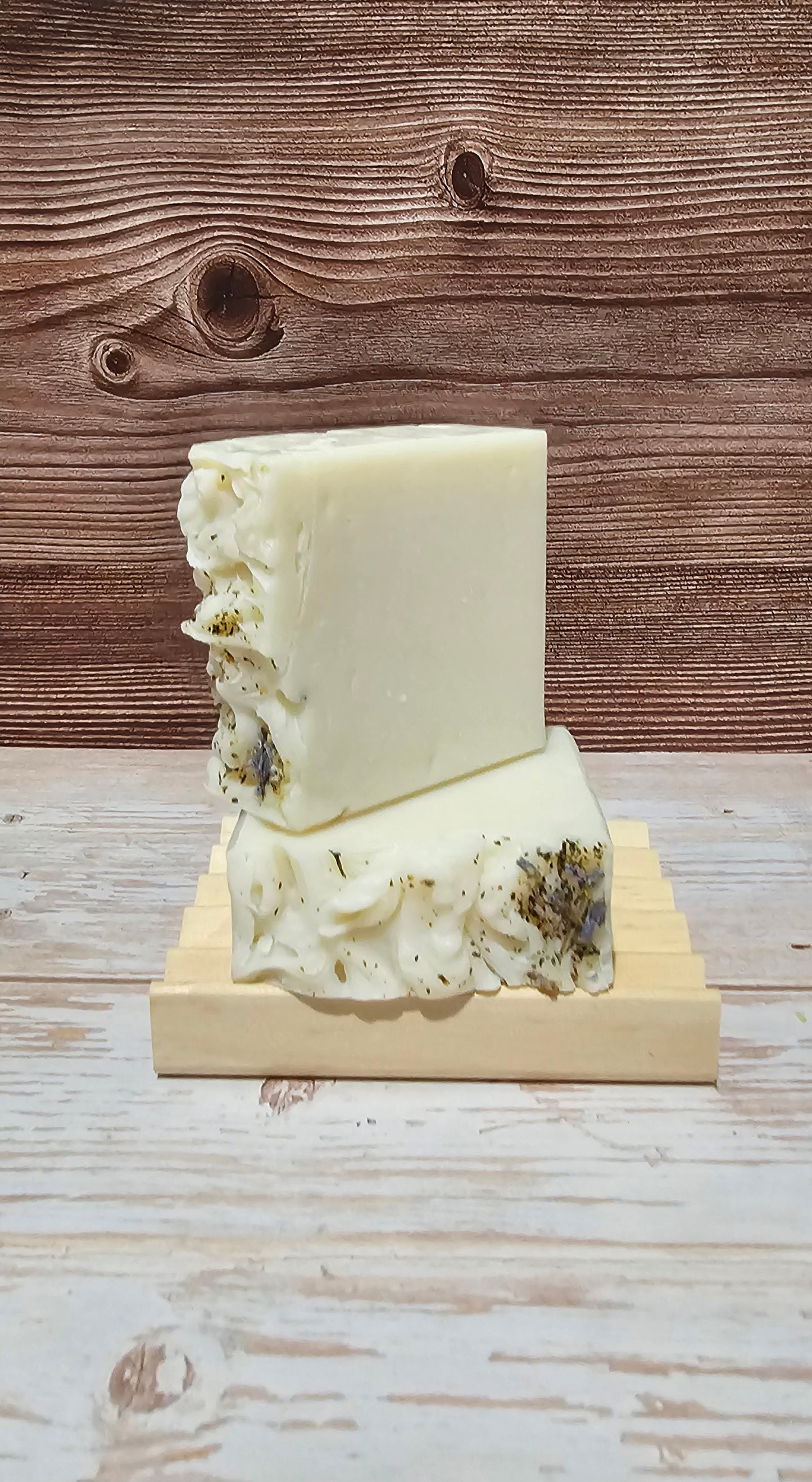 Two bars of white soap with green herbs on a wooden stand against a wooden background