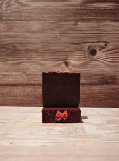 A dark red wax melt or soap block displayed in an open wooden gift box with a small red bow on the front, set against a rustic wooden plank background.