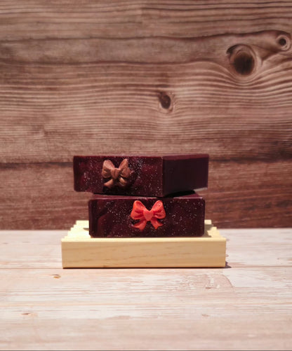 Two small rectangular boxes stacked on a light wood base, set against a rustic wooden plank background. The top box is dark brown with a glittery gold bow, and the bottom box is deep red with a glittery red bow.