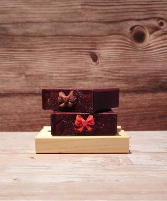 Two small rectangular boxes stacked on a light wood base, set against a rustic wooden plank background. The top box is dark brown with a glittery gold bow, and the bottom box is deep red with a glittery red bow.