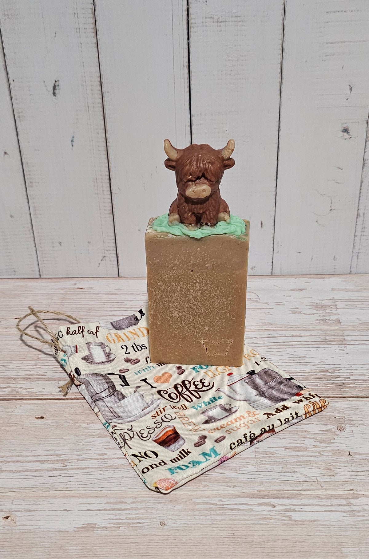 A rectangular bar of brown soap with a small Highland cow figurine sitting on top, displayed on a patterned coffee-themed cloth against a white wood plank background.