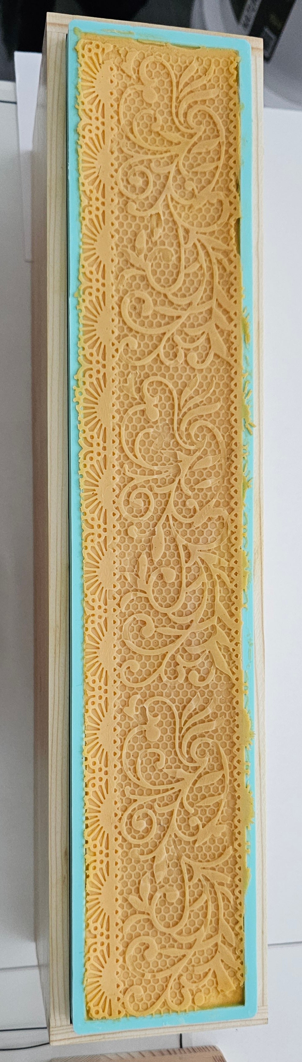 A rectangular piece of textured yellow lace or embroidered fabric with an ornate floral pattern, bordered by a light blue inner edge and a beige outer frame, shown against a white background.