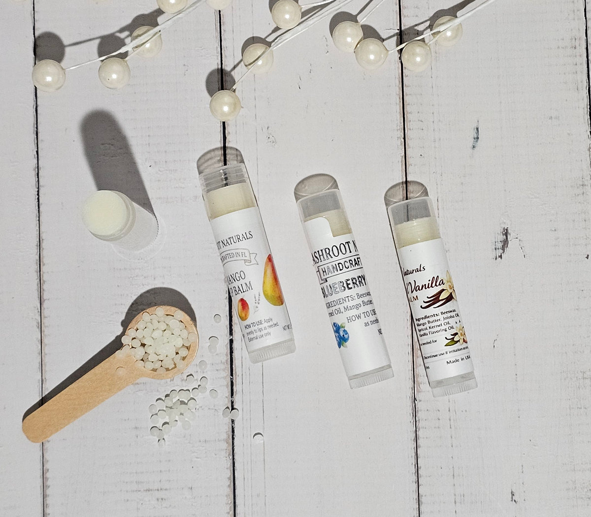 Three lip balm tubes labeled Planty Balms, Shroott Handmade, and Mandel's Vanilla, arranged on a white wooden surface with a wooden scoop of balm beads and decorative pearl garlands.