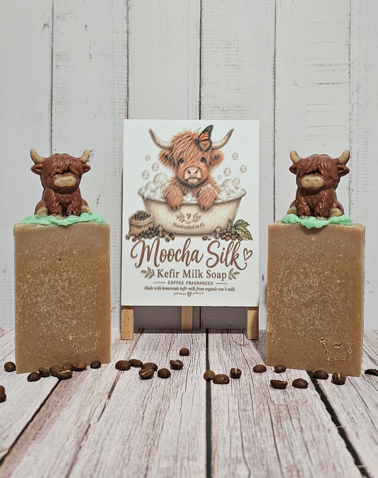 Two rectangular bars of Moocha Silk Kefir Milk Soap stand on a rustic wood surface, each topped with a small brown cow figurine sitting on green leaves. A centered product card with an illustration of a shaggy cow in a bathtub reads 'Moocha Silk Kefir Milk Soap' and includes the brand name Moocha Silk.