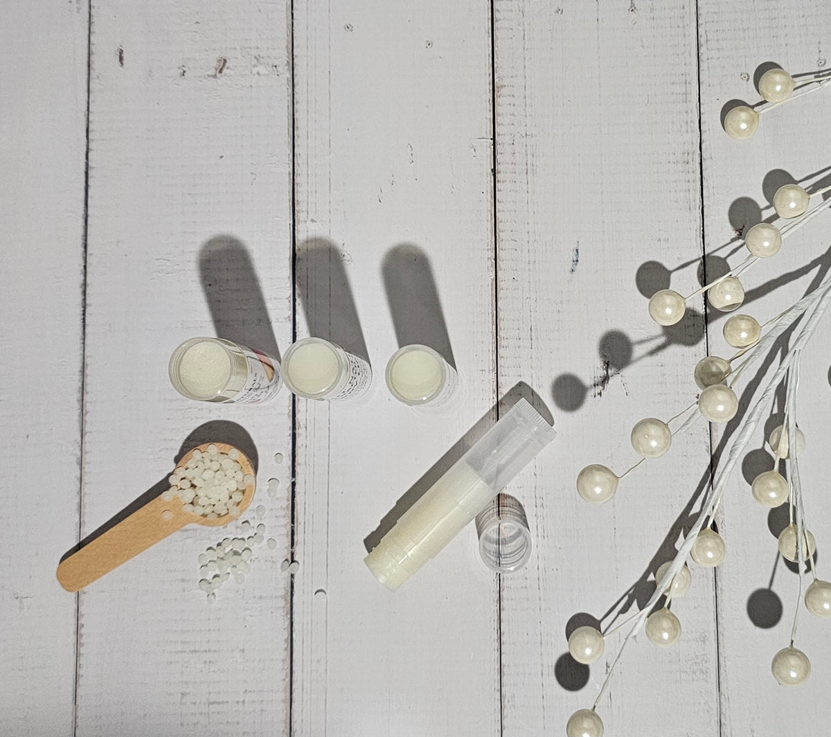 A styled flat-lay of lip balm products on a white wooden background, including three small jars, a clear tube of white balm, loose balm pieces in a wooden scoop, and decorative pearl branches casting shadows.
