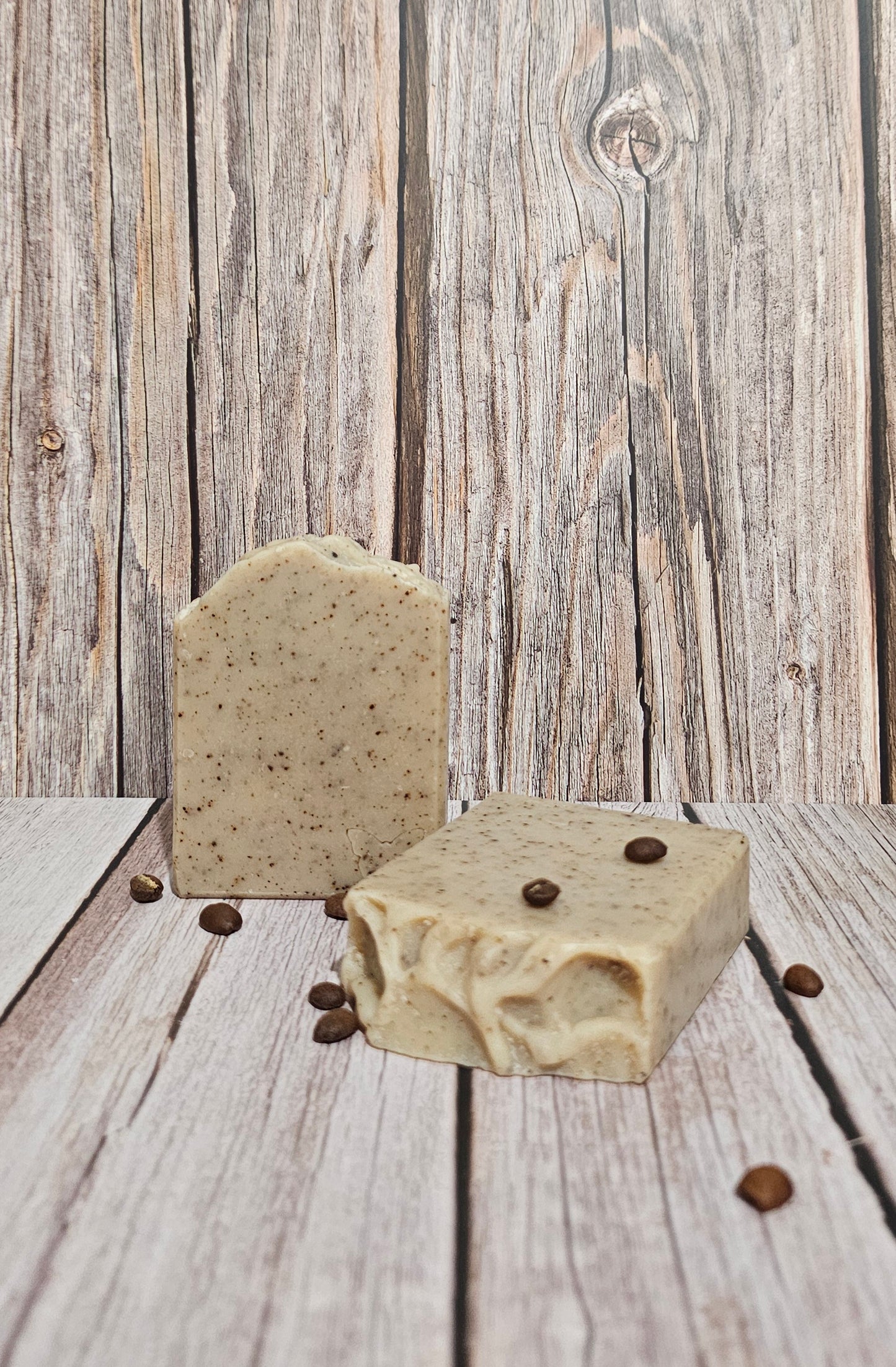 Two rectangular bars of handmade soap with a speckled oatmeal texture and scattered coffee beans, set against a rustic gray wooden plank background.