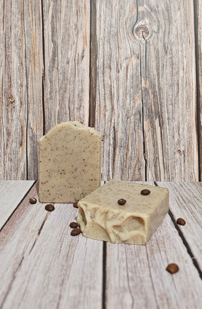Two rectangular bars of handmade soap with a speckled oatmeal texture and scattered coffee beans, set against a rustic gray wooden plank background.