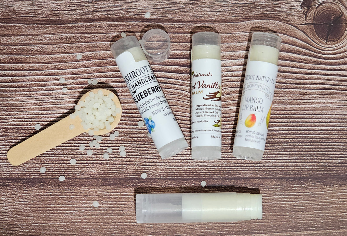 Four lip balm tubes and a small wooden scoop filled with white balm pieces are arranged on a brown wood-grain surface. The labels show flavors including Blueberry, Vanilla, and Mango.
