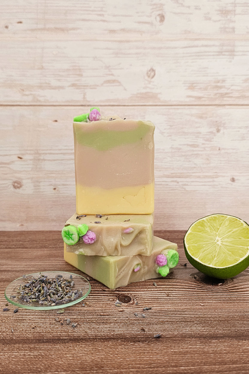 Three stacked bars of handmade soap on a rustic wooden surface, with a small dish of dried lavender and a lime half beside them. The soap has a two-tone beige and pale yellow top bar, and light green and pale yellow lower bars decorated with small pink and green flower pieces.