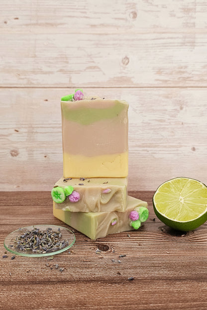 Three stacked bars of handmade soap on a rustic wooden surface, with a small dish of dried lavender and a lime half beside them. The soap has a two-tone beige and pale yellow top bar, and light green and pale yellow lower bars decorated with small pink and green flower pieces.
