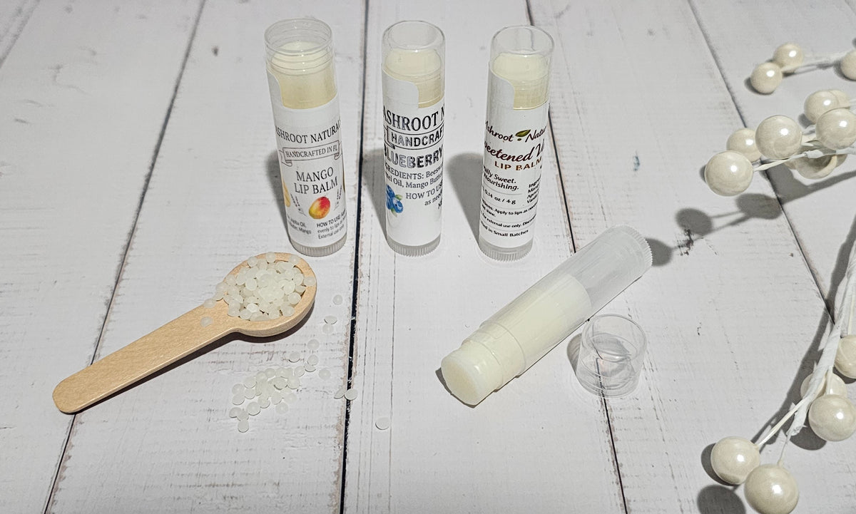 Three tubes of lip balm and one opened tube with its cap beside a small wooden spoon of balm pieces, arranged on a white wooden surface with decorative pearl beads.