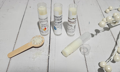 Three tubes of lip balm and one opened tube with its cap beside a small wooden spoon of balm pieces, arranged on a white wooden surface with decorative pearl beads.