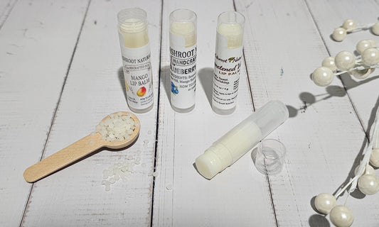 Three tubes of lip balm and one opened tube with its cap beside a small wooden spoon of balm pieces, arranged on a white wooden surface with decorative pearl beads.