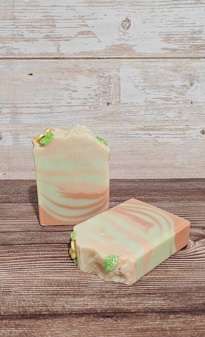 Two handmade soap bars with a light green and peach swirl pattern, decorated with small green leaf inclusions, displayed on a rustic wooden surface.