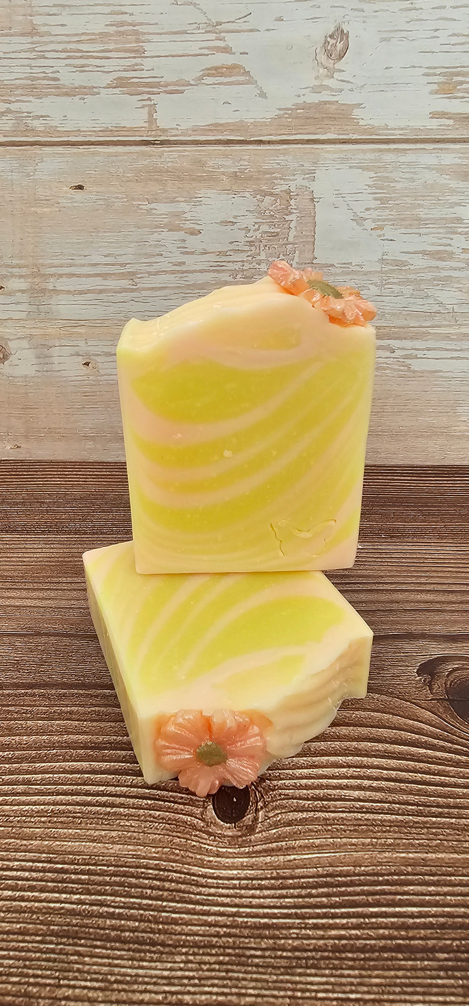 Two rectangular bars of pale yellow and white marbled soap with small pink flower decorations on top, arranged on a rustic wooden surface.