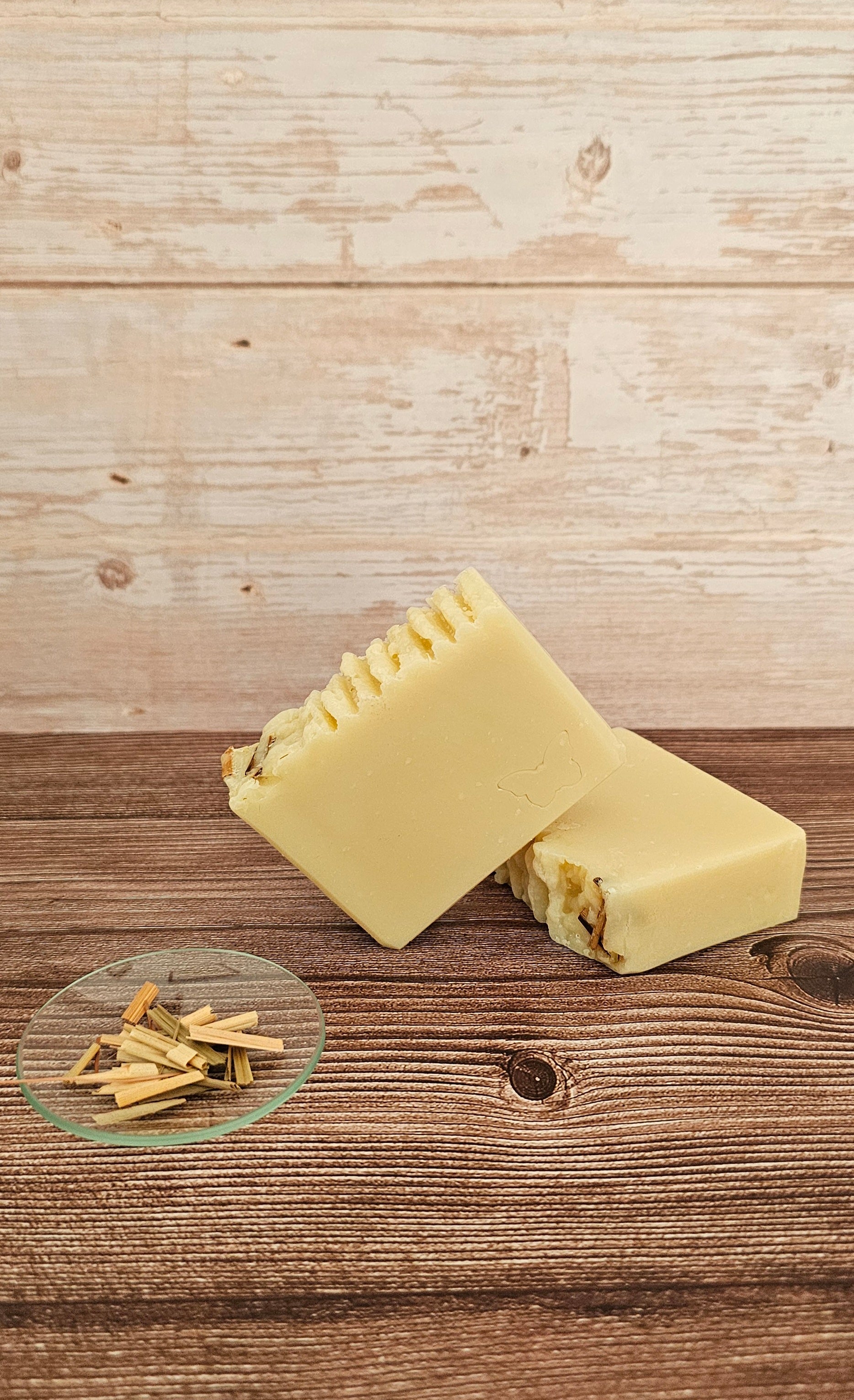 Two pale yellow handmade soap bars with textured tops and visible natural filler pieces, arranged on a rustic wooden surface with a small glass dish of dried botanicals in the foreground, against a whitewashed wood plank backdrop.