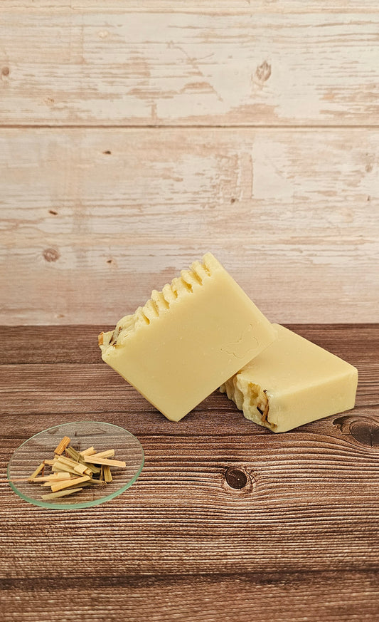 Two pale yellow handmade soap bars with textured tops and visible natural filler pieces, arranged on a rustic wooden surface with a small glass dish of dried botanicals in the foreground, against a whitewashed wood plank backdrop.